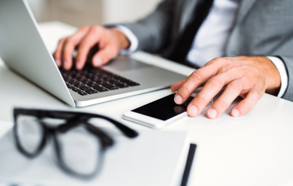 A midsection of businessman with smartphone, glasses and laptop sitting at the table in an office, working. Copy space.