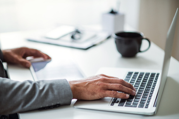 A midsection of businessman with tablet and laptop sitting at the table in an office, working. Copy space.