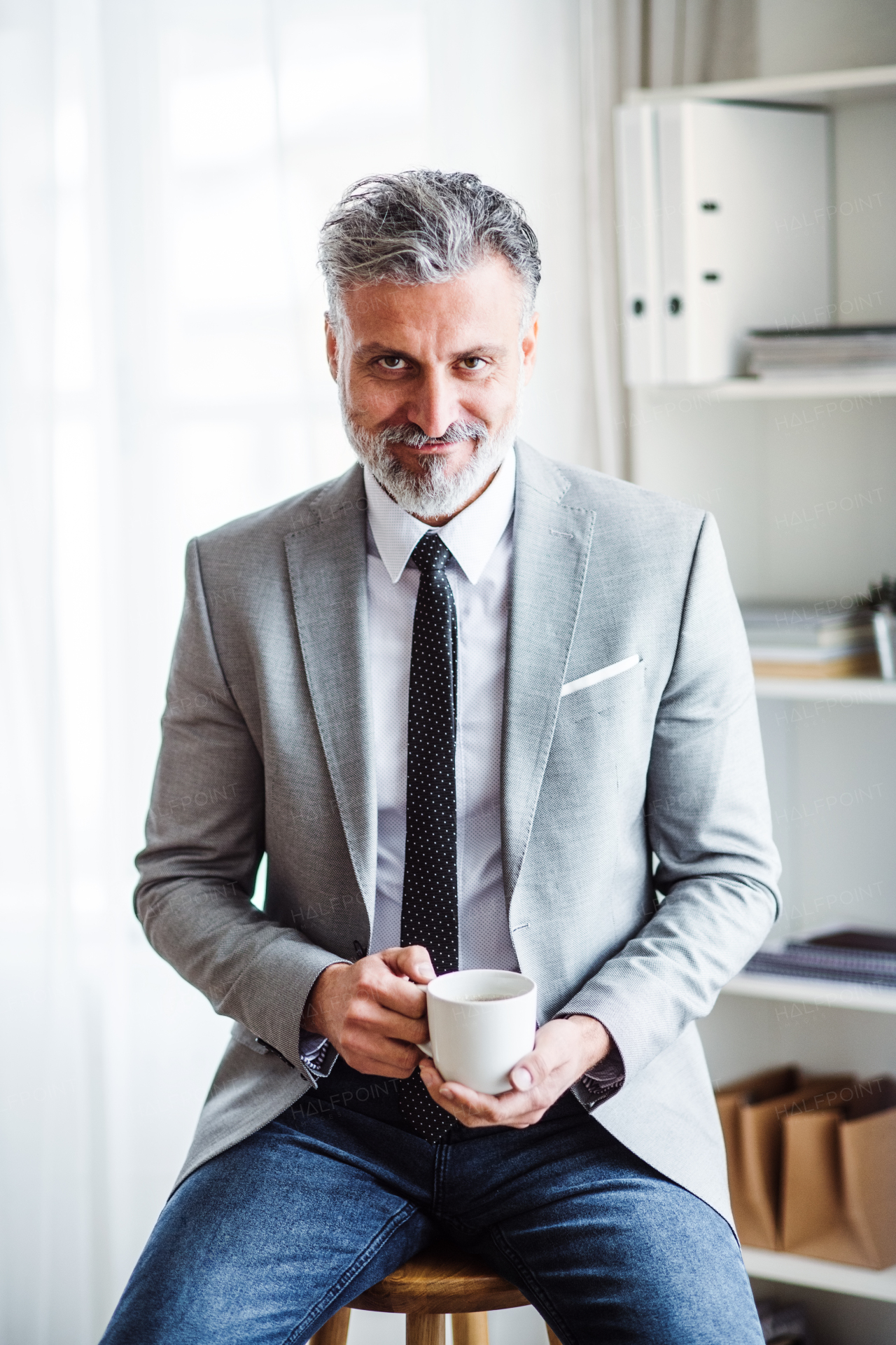 A mature businessman sitting in an office on a stool, holding a cup of coffee.