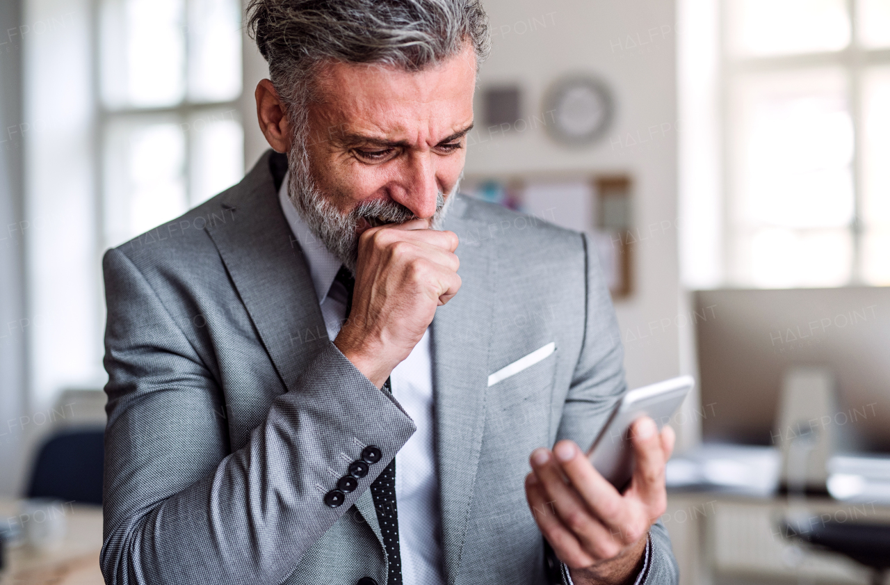 A sad and frustrated businessman with smartphone standing in an office, reading bad news.