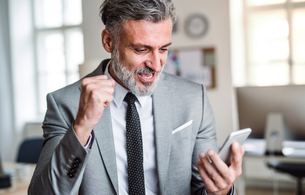 A mature businessman with smartphone standing in an office, expressing excitement.