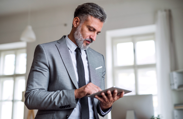 A waist-up portrait of mature businessman standing in an office, using tablet.