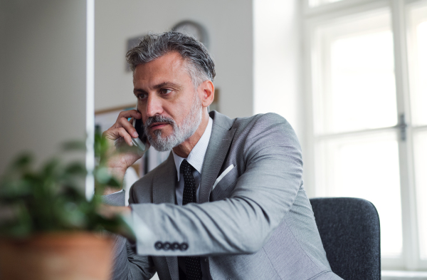 Serious mature businessman with smartphone and desktop computer sitting at the table in an office, making a phone call.