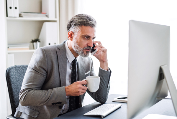 Serious mature businessman with smartphone, coffee and desktop computer sitting at the table in an office, making a phone call.