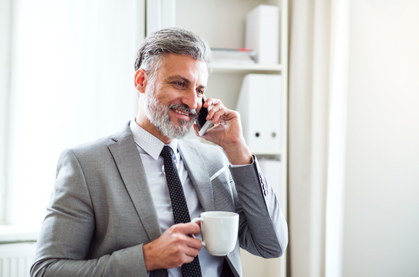 Mature businessman with smartphone and coffee standing in an office, making a phone call.