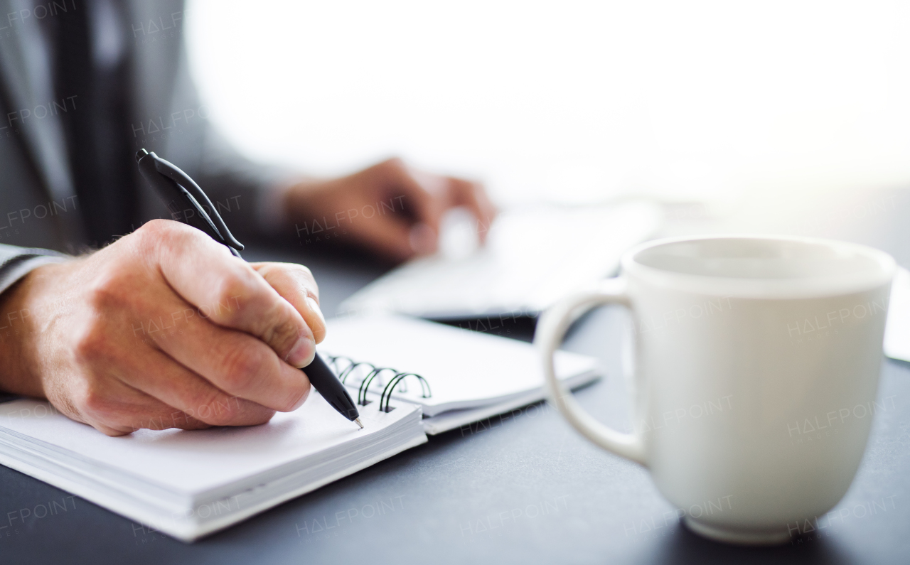 A midsection of businessman with cup sitting at the table in an office, making notes.