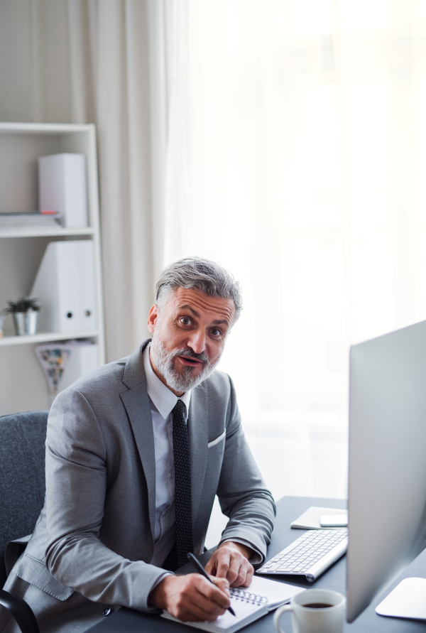 A businessman with desktop computer sitting at the table in an office, making notes.