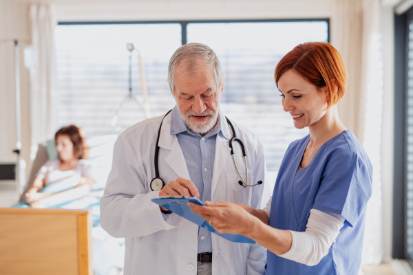 Portrait of senior male doctor standing in hospital room, talking to a female nurse.