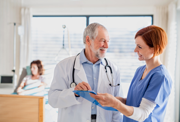 Portrait of senior male doctor standing in hospital room, talking to a female nurse.