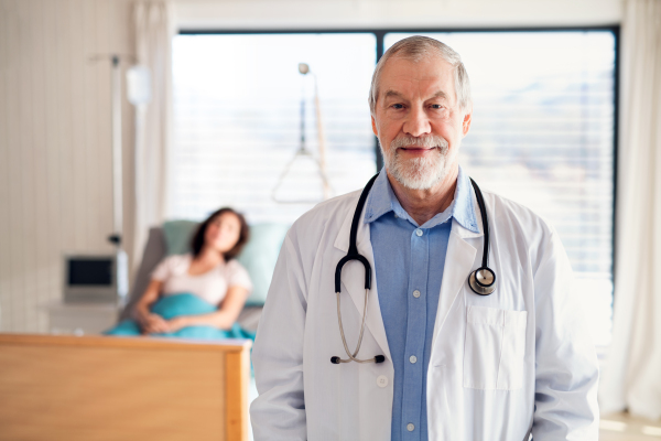 A portrait of senior male doctor standing in hospital room, patient in the background.