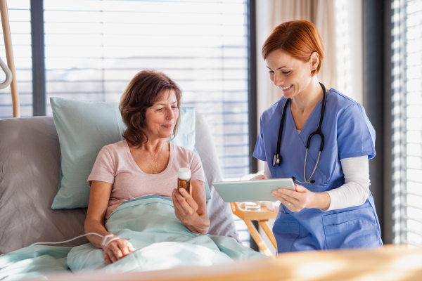 A front view of female doctor with tablet talking to patient in bed in hospital.