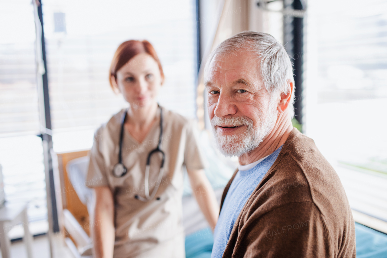 A senior man patient and doctor sitting on bed in hospital, looking at camera.