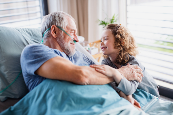 A cheerful small girl visiting ill grandfather in bed in hospital room.