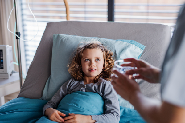 Unrecognizable doctor or nurse giving glass of water to a small girl in bed in hospital.