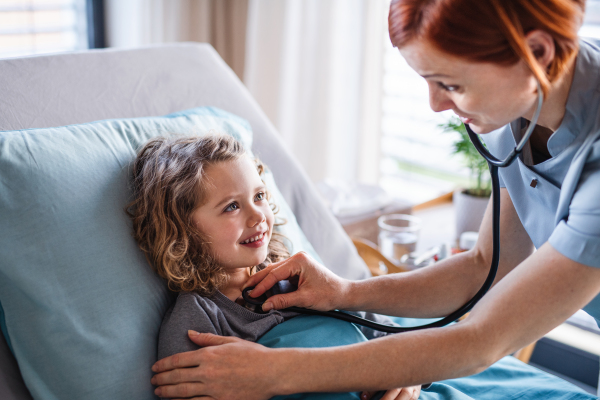 Friendly female doctor with stethoscope examining small girl in bed in hospital.