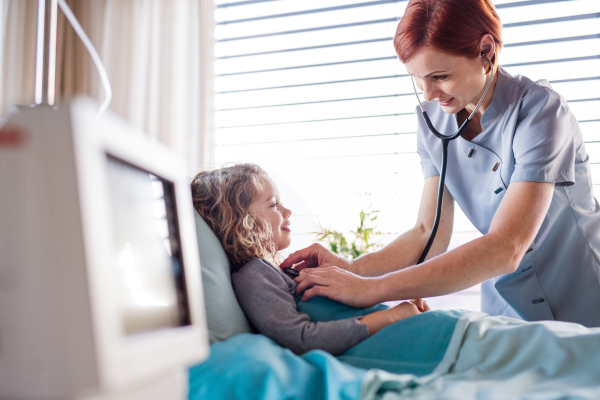 Friendly female doctor with stethoscope examining small girl in bed in hospital.