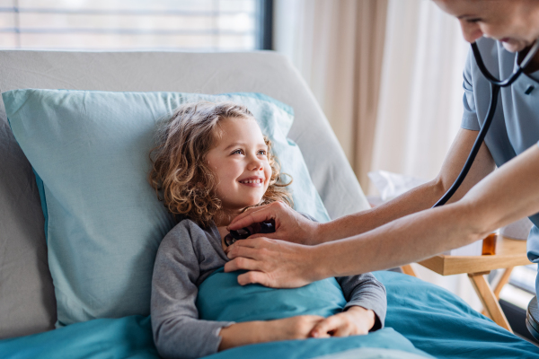 Friendly female doctor with stethoscope examining small girl in bed in hospital, midsection.