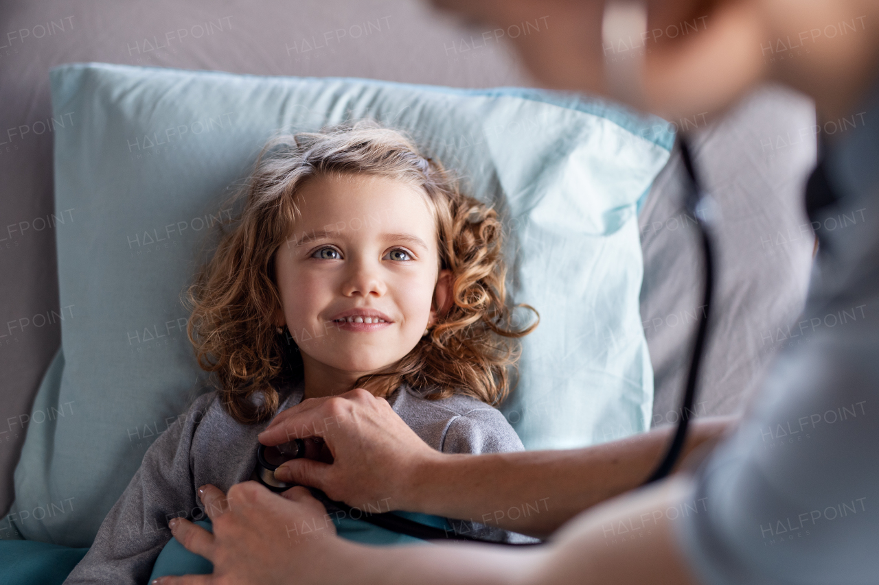 Unrecognizable female doctor with stethoscope examining small girl in bed in hospital.