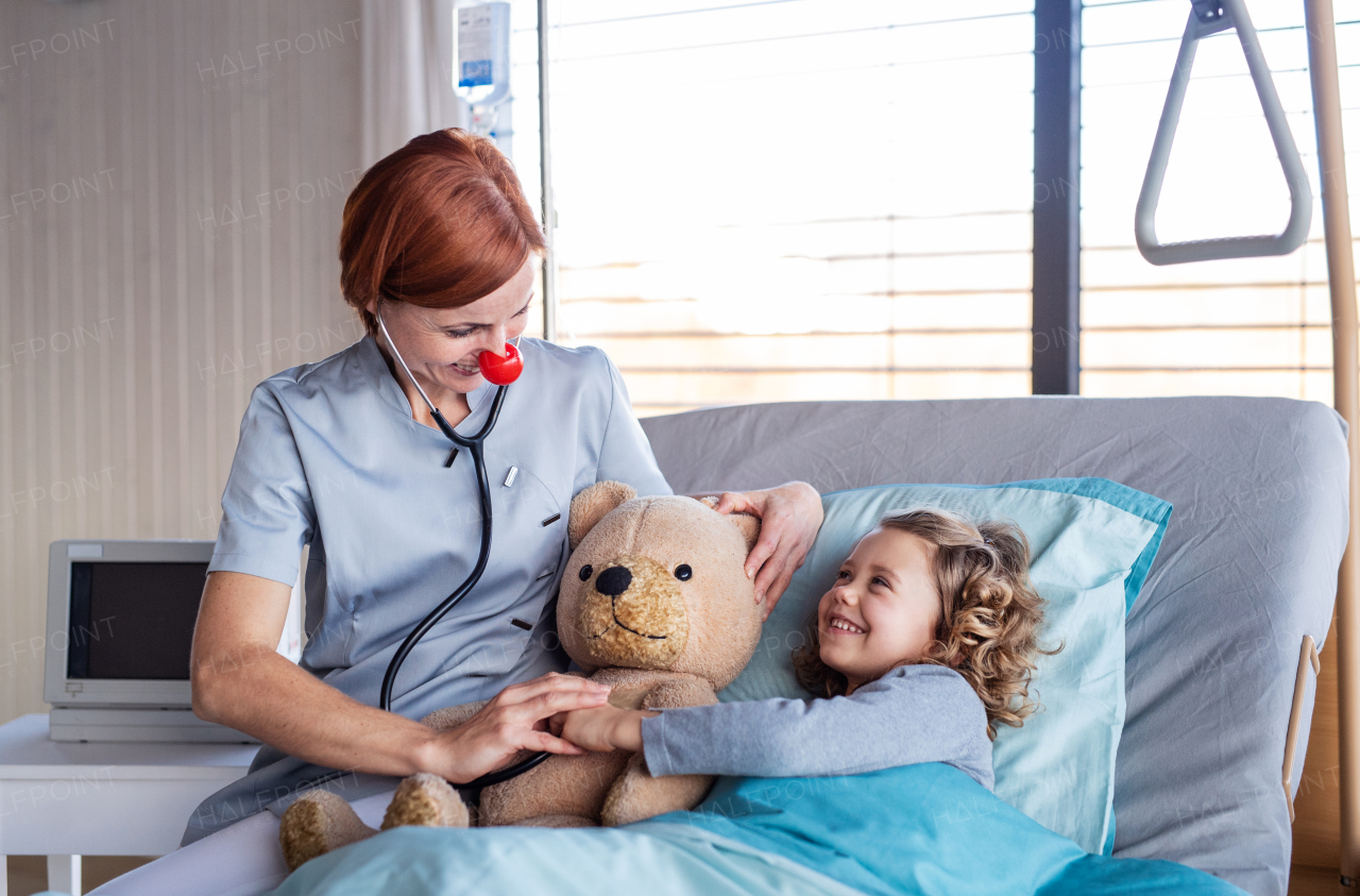 Friendly female doctor with stethoscope examining small girl in bed in hospital.