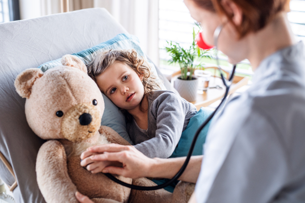 Friendly female doctor with stethoscope examining small girl in bed in hospital.