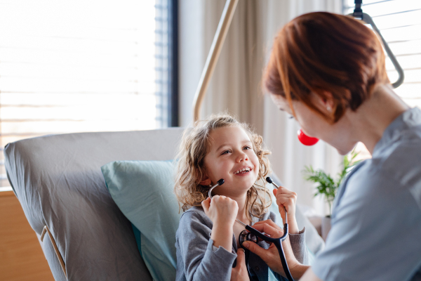 Friendly female doctor with stethoscope examining small girl in bed in hospital.