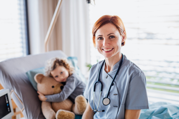 Friendly female doctor and small girl in bed in hospital, looking at camera.