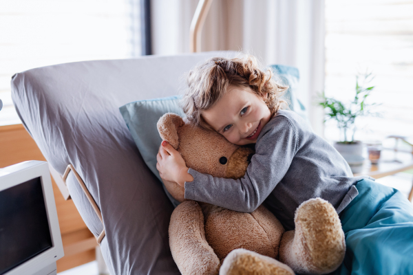 Smiling small girl with teddy bear in bed in hospital, looking at camera.