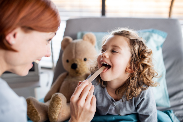 Female doctor with tongue depressor examining small girl in bed in hospital.