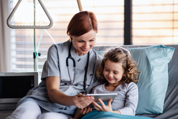 Front view of friendly female doctor with tablet and small girl in bed in hospital.