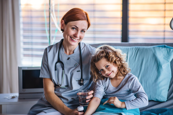 Friendly female doctor with stethoscope examining small girl in bed in hospital.