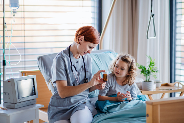 Friendly female doctor with medication examining small girl in bed in hospital.