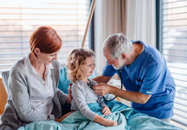 Senior doctor with stethoscope examining a small hospitalized girl with mother in hospital.