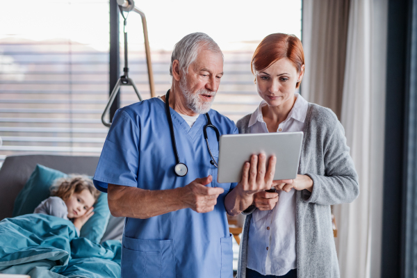 Senior doctor with tablet talking to mother of small hospitalized girl in hospital.