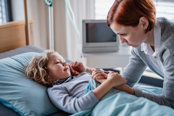 Caring mother visiting small girl daughter in bed in hospital, stroking her face.