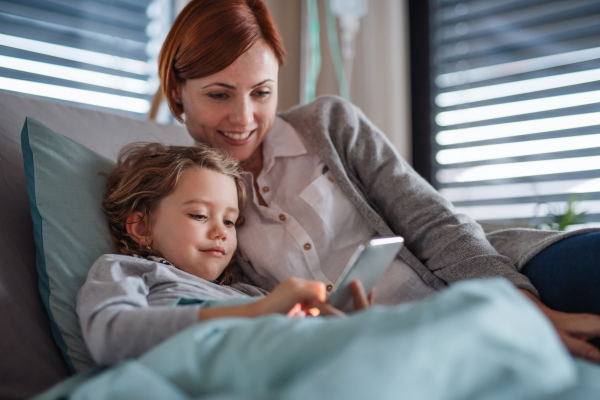 Happy small girl with mother in bed in hospital, using smartphone to pass time.