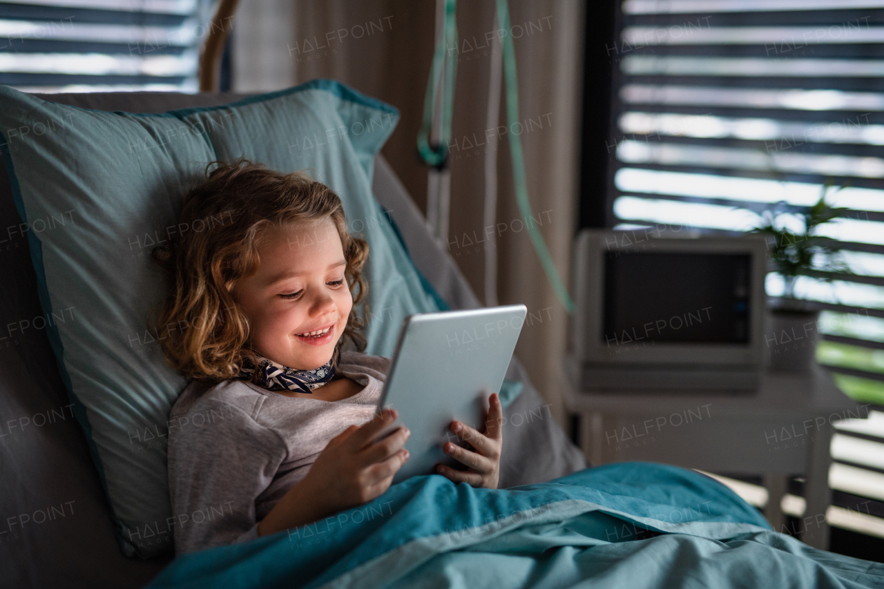 Bored small girl lying in bed in hospital, using tablet.