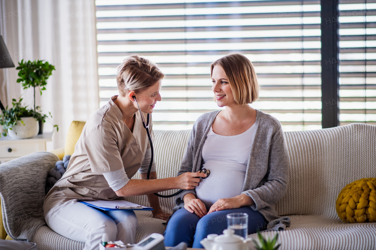 A healthcare worker with stethoscope examining pregnant woman indoors at home.