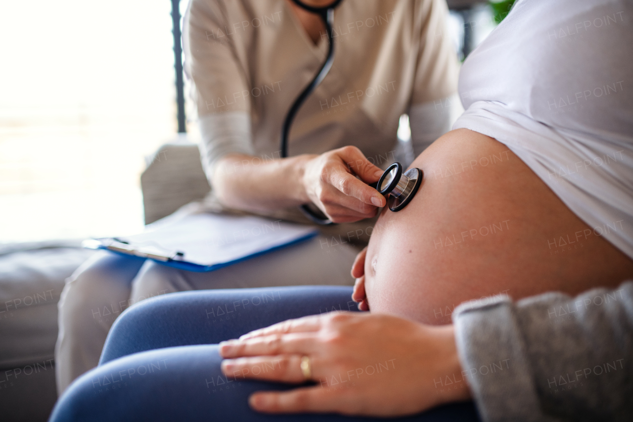 A midsection of unrecognizable healthcare worker examining pregnant woman indoors at home.