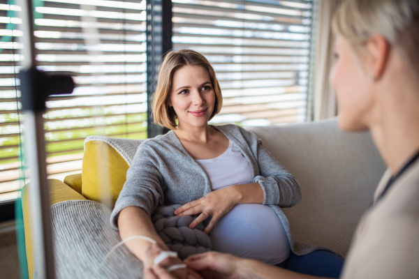 Unrecognizable healthcare worker with IV drip examining pregnant woman indoors at home.