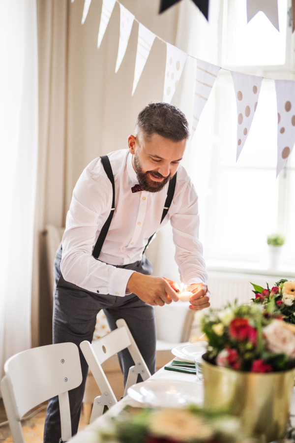 A portrait of hipster mature man indoors in a room set for a party, lighting candles.