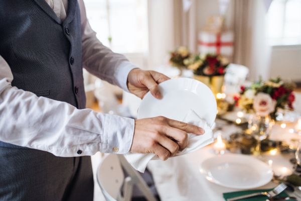 A midsection of unrecognizable man indoors in a room set for a party, polishing plates.