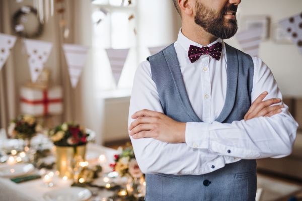 A midsection of a hipster mature man standing indoors in a room set for a party, arms crossed.