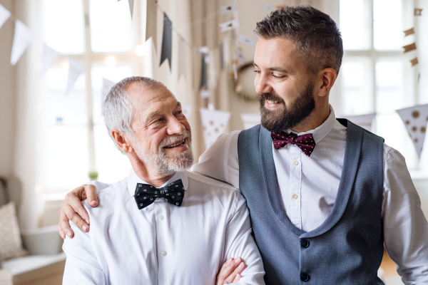 A portrait of a senior and hipster mature man standing indoors in a room set for a party, looking at each other.