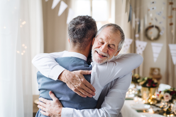 A portrait of a senior and hipster mature man standing indoors in a room set for a party, hugging.