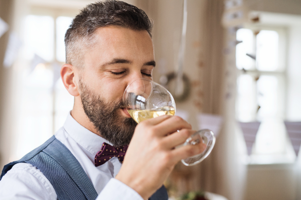 A portrait of hipster mature man indoors in a room set for a party, drinking white wine.