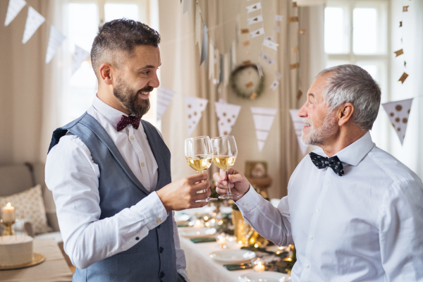 A senior and mature man standing indoors in a room set for a party, clinking glasses with wine.