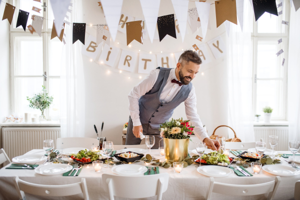 A portrait of hipster mature man indoors in a room set for a party, setting table.