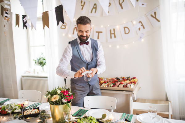 A mature man with bow and vest setting a table for an indoor party, polishing a glass.