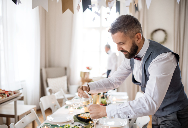 A mature man with bow and vest setting a table for an indoor party, polishing a glass.