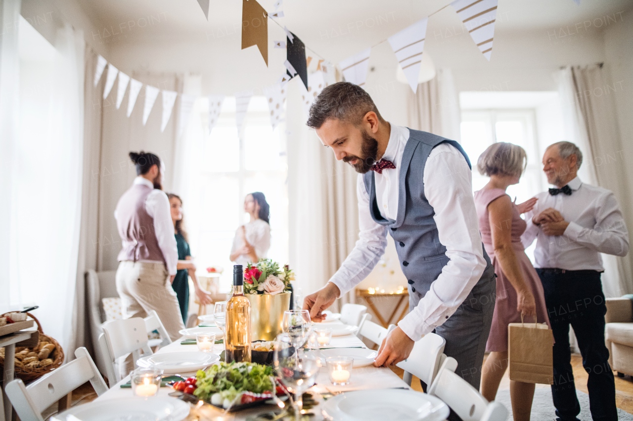 A mature man with bow and vest setting a table for an indoor party, guests in the background.